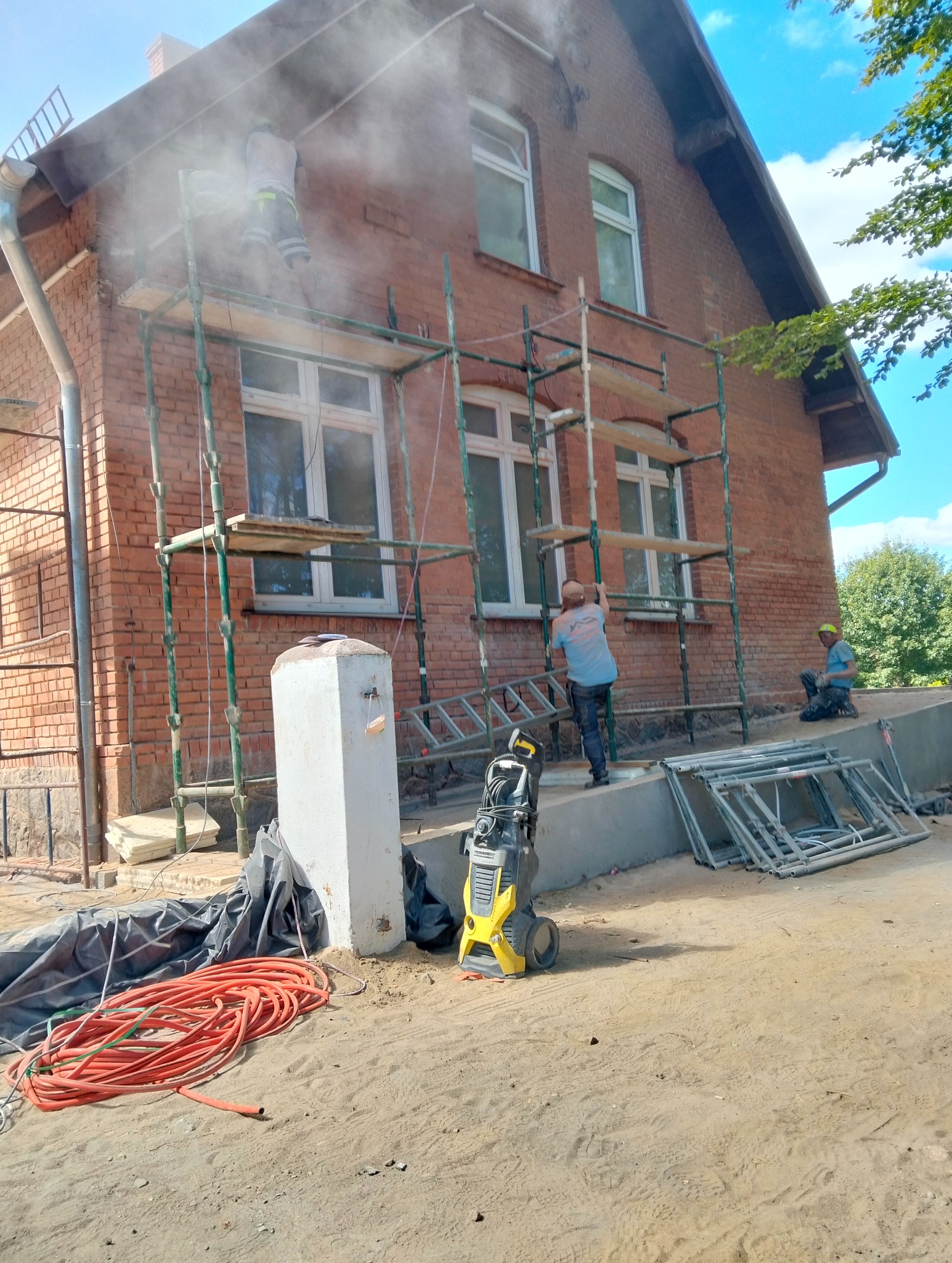 Czyszczenie ceglanej elewacji domu z rusztowania za pomocą myjki ciśnieniowej. Widoczne węże, drabina i elementy rusztowania. Prace na zewnątrz budynku.