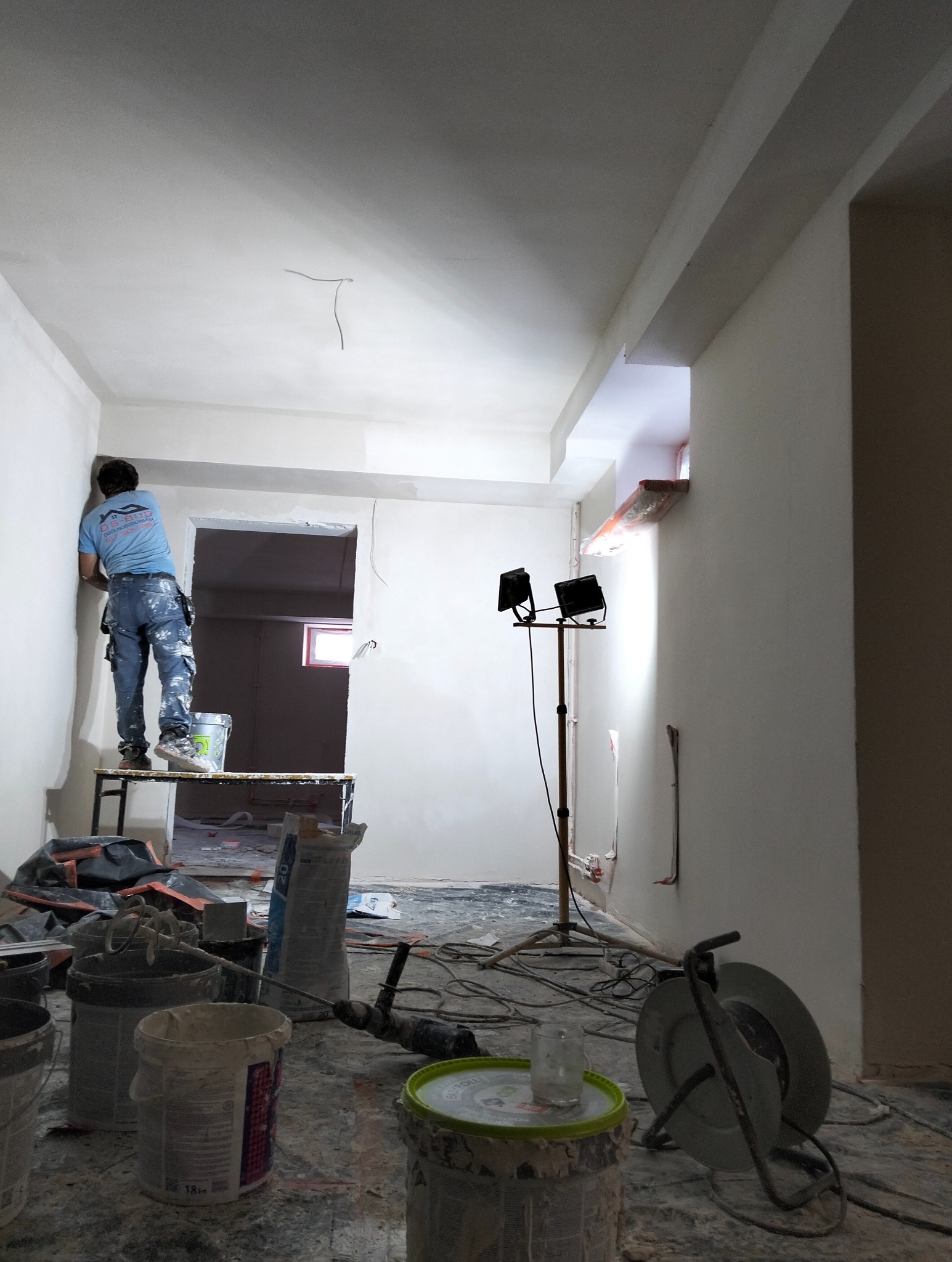 Pracownik na podeście szpachluje ścianę w jasnym wnętrzu. Widoczne narzędzia, oświetlenie budowlane i surowe wykończenie. Ujęcie z dołu, podkreślające etap prac.
