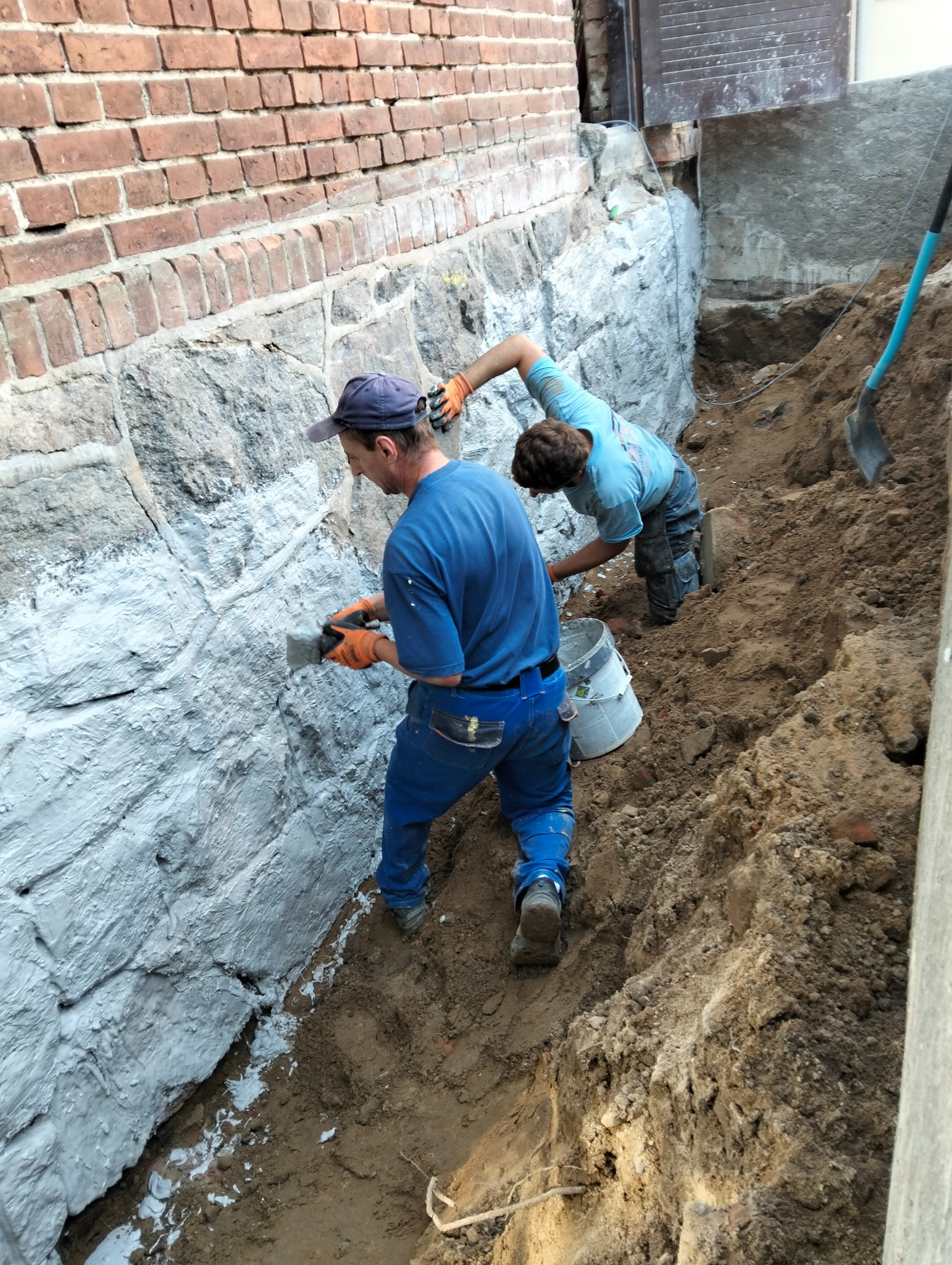 Dwóch robotników zabezpiecza fundamenty budynku, nakładając szpachelką szarą warstwę izolacyjną na kamienną ścianę. Wykop ziemny, ceglany mur w tle.