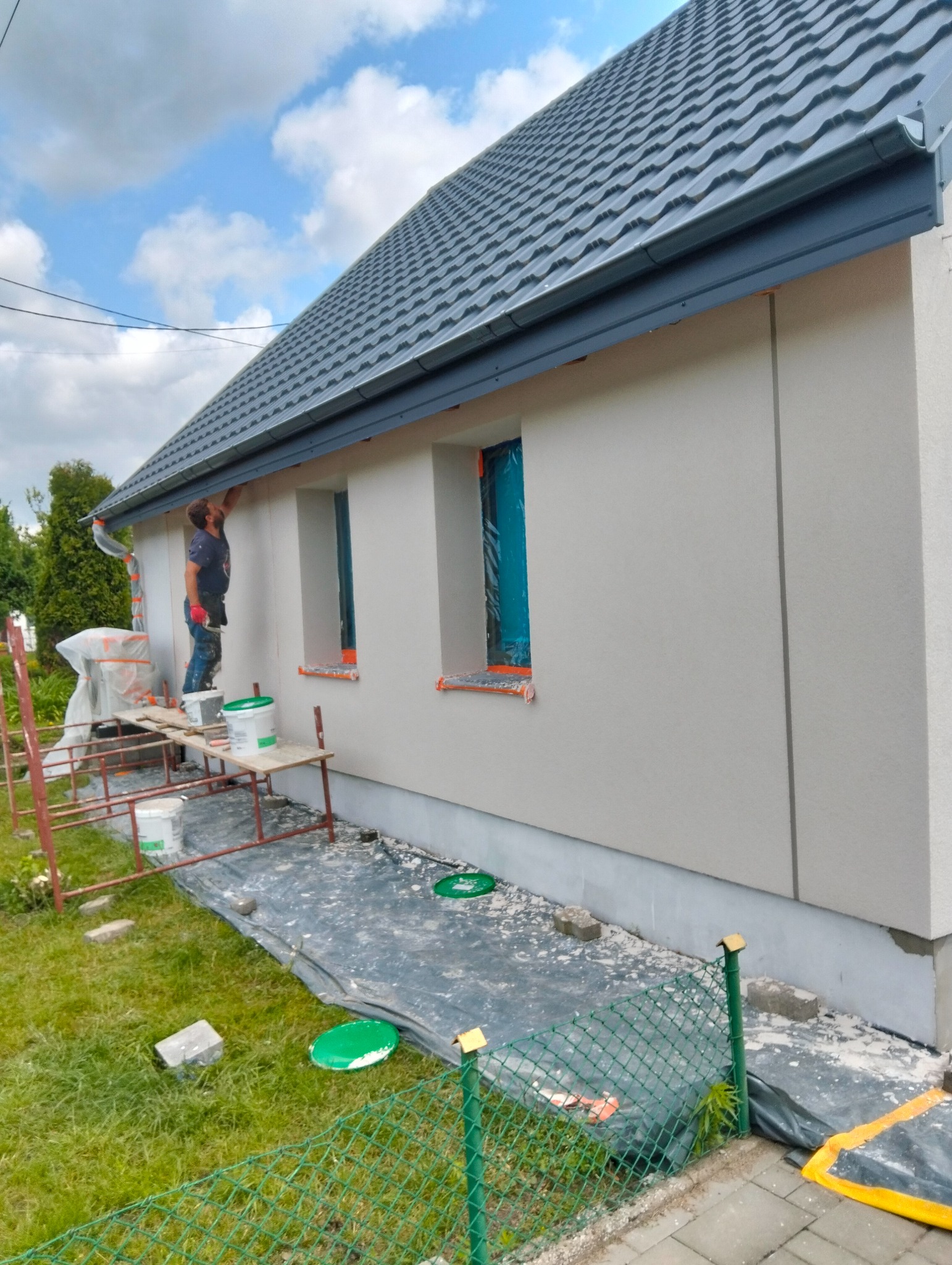 Pracownik maluje elewację domu jednorodzinnego na zewnątrz. Widoczne rusztowanie, puszki z farbą i zabezpieczone okna folią. Szara dachówka i jasna elewacja.