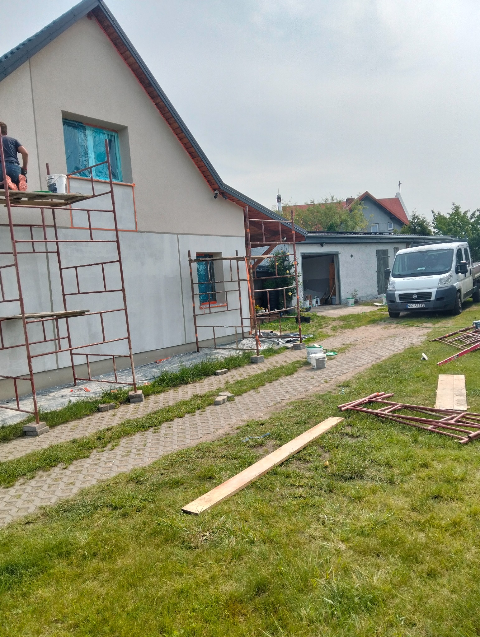 Dom jednorodzinny w trakcie prac elewacyjnych. Widoczne rusztowania, częściowo otynkowana ściana, zabezpieczone okna folią. Na podjeździe zaparkowany biały samochód dostawczy.