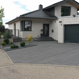 Eukor Okna & Drzwi - Nowoczesny dom z garażem i czarną bramą garażową, widoczny z zewnątrz. Szara kostka brukowa na podjeździe. Minimalistyczny styl, jasna elewacja i ciemne okna.