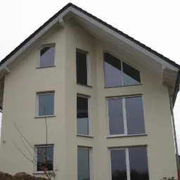 Dwukondygnacyjny dom z beżową elewacją i oknami o różnych kształtach, w tym trójkątnym na poddaszu. Widok z dołu, pochmurne niebo.