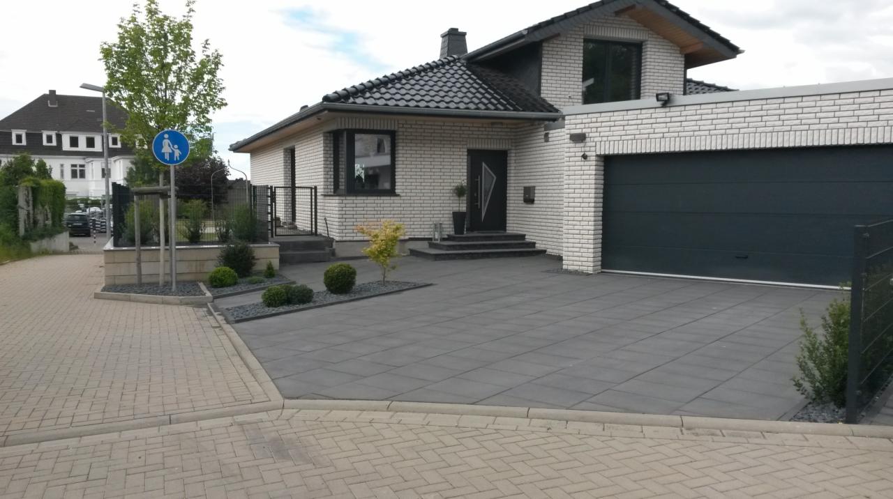 Nowoczesny dom z garażem i czarną bramą garażową, widoczny z zewnątrz. Szara kostka brukowa na podjeździe. Minimalistyczny styl, jasna elewacja i ciemne okna.