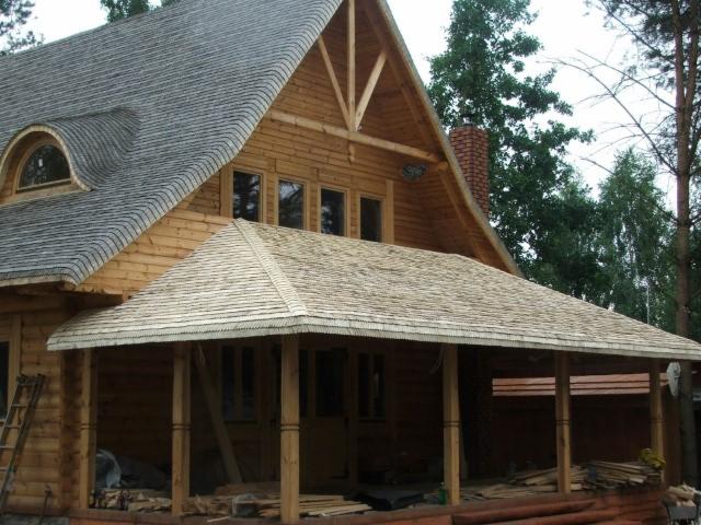 Drewniany dom z dachem pokrytym wiórem osikowym, widoczna weranda z drewnianymi słupami i fragment dachu głównego. Na pierwszym planie porozrzucane deski i drabina oparta o ścianę.