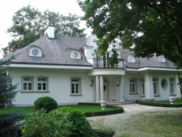 Elegancki, biały dom jednorodzinny z dachem pokrytym szarym wiórem osikowym i okrągłymi oknami na poddaszu, otoczony zadbanym ogrodem.