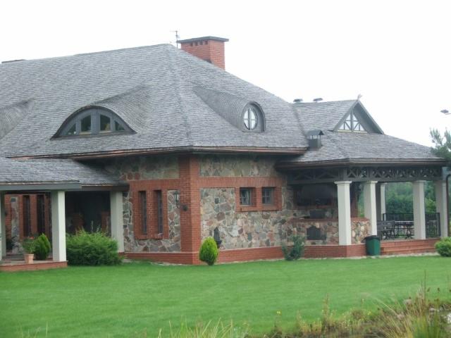 Dom jednorodzinny z dachem pokrytym szarym gontem bitumicznym, okna dachowe w kształcie półksiężyca, kamienno-ceglana elewacja i zadaszona weranda z kolumnami.