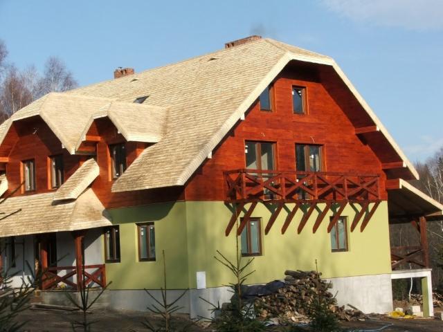 Dom z dachem krytym wiórem osikowym, widok z zewnątrz, drewniane elementy konstrukcyjne, zielona elewacja, stos drewna przy ścianie.