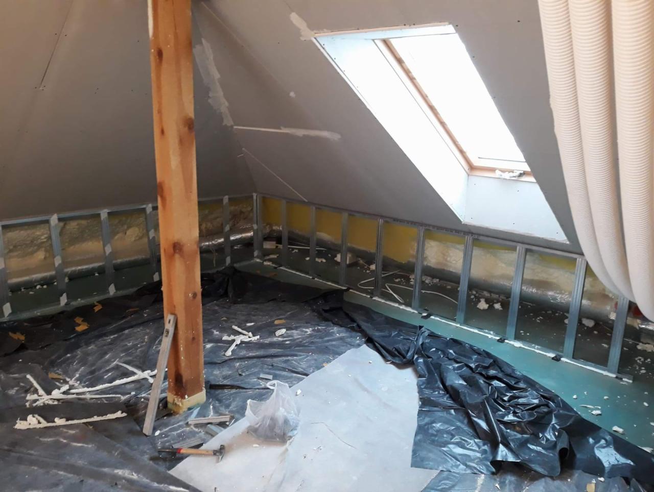 Poddasze w trakcie adaptacji: metalowa konstrukcja ścian działowych z ociepleniem, okno dachowe, przewody wentylacyjne, zabezpieczone podłogi folią budowlaną, widoczny poziomica i młotek.