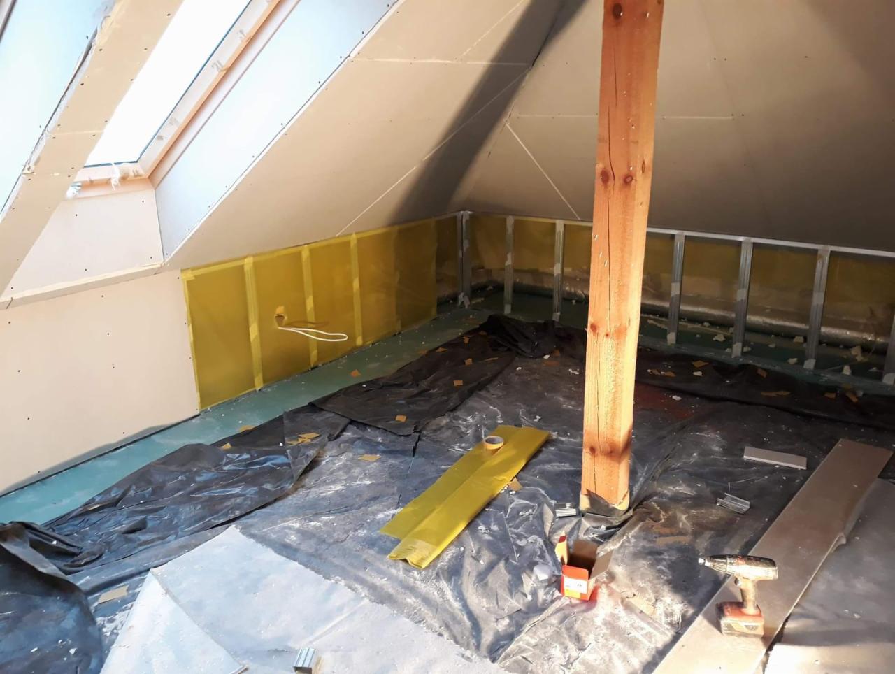 Adaptacja poddasza: widok na ocieplone żółtą wełną ściany, metalowy stelaż, drewniany słup nośny, rozłożoną folię ochronną, narzędzia i materiały budowlane w trakcie prac wykończeniowych.