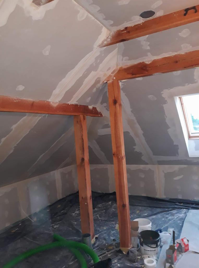 Skośne poddasze w trakcie wykańczania płytami gipsowo-kartonowymi, widoczne drewniane belki konstrukcyjne, szpachlowane łączenia płyt, okno dachowe i czarna folia ochronna na podłodze.