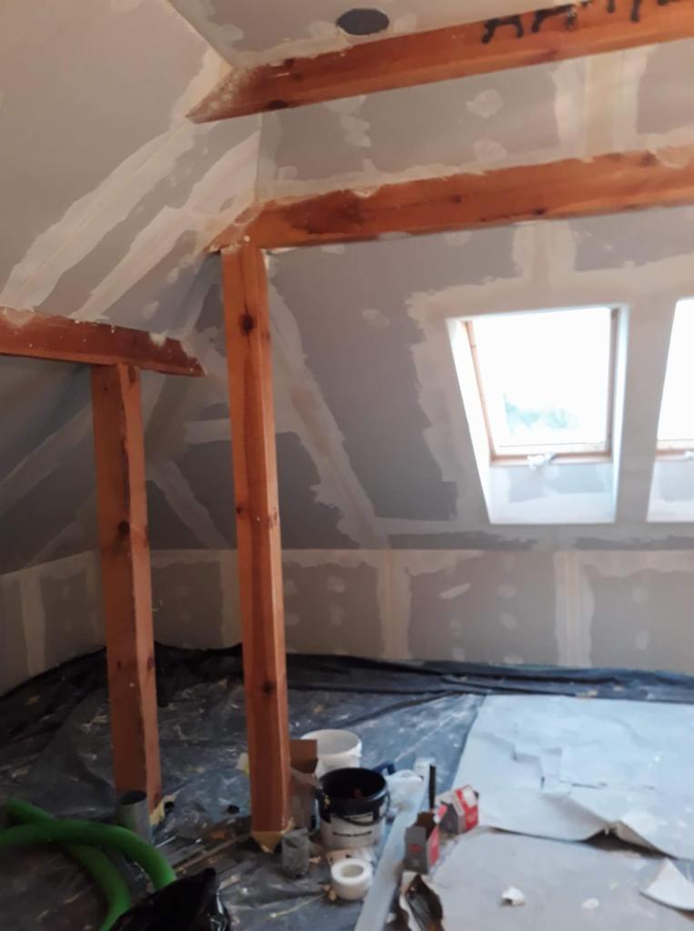 Poddasze w trakcie remontu: widoczne drewniane belki konstrukcyjne, ściany pokryte płytami gipsowo-kartonowymi z widocznymi łączeniami, folia ochronna na podłodze i narzędzia malarskie.