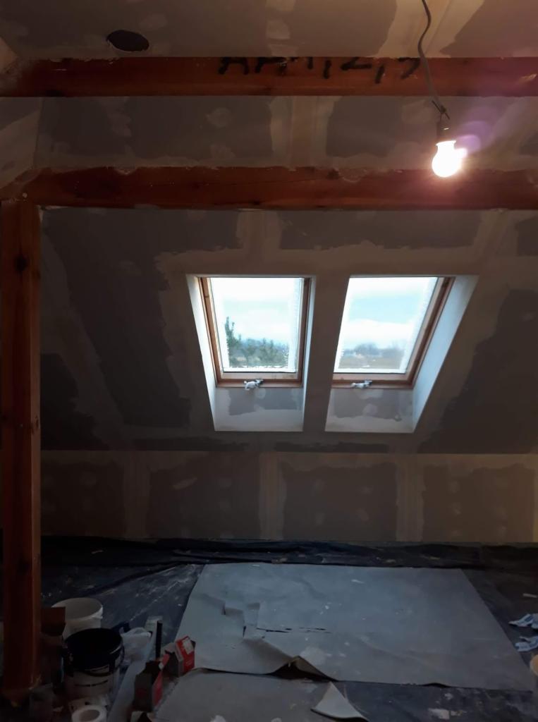 Poddasze w trakcie remontu, widoczne dwa okna dachowe, ściany pokryte płytami gipsowo-kartonowymi, sufit w trakcie szpachlowania, na podłodze folia ochronna i resztki materiałów budowlanych, surowa...