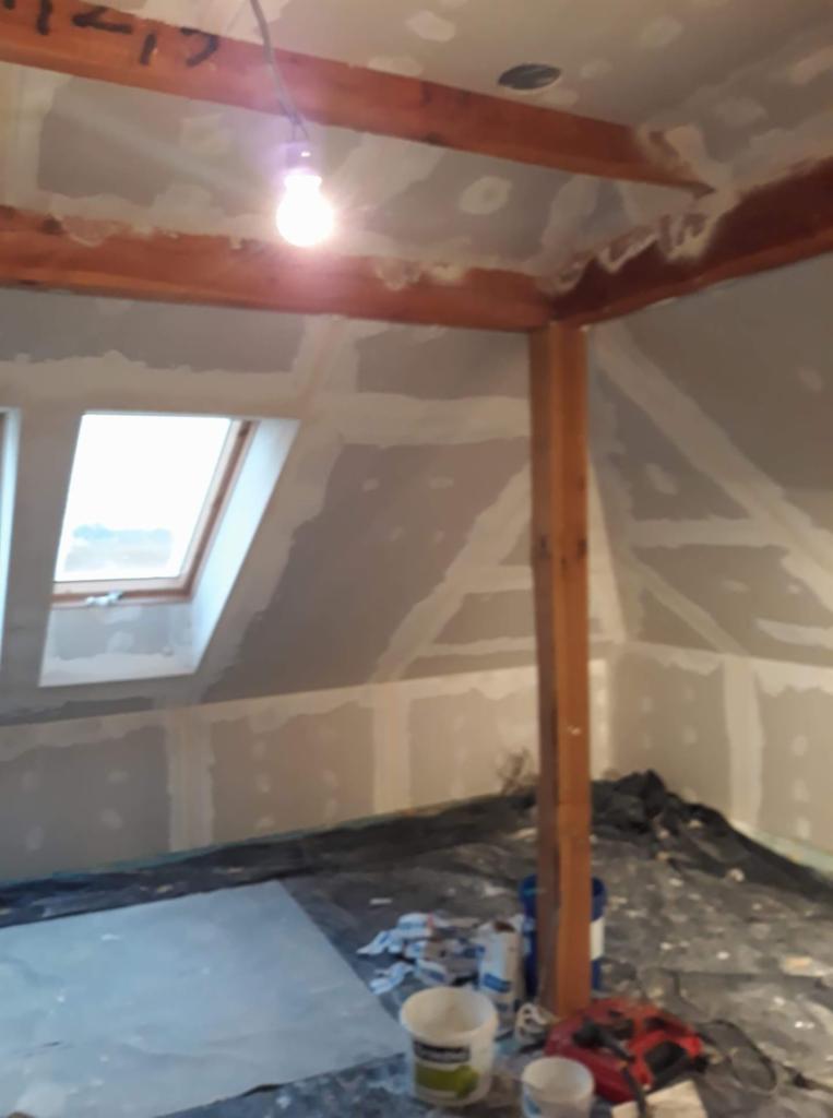 Poddasze w trakcie wykańczania, widoczne płyty gipsowo-kartonowe na ścianach i suficie, drewniana konstrukcja nośna, okno dachowe i tymczasowe oświetlenie żarówką.