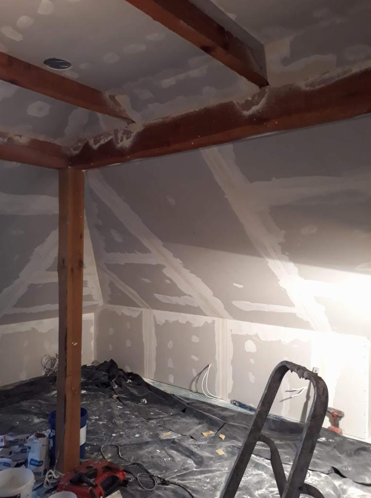 Poddasze w trakcie remontu, widoczne płyty gipsowo-kartonowe na ścianach i suficie, szpachlowane łączenia, drewniane belki konstrukcyjne, folia ochronna na podłodze i narzędzia.