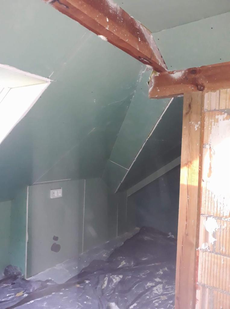 Poddasze w trakcie wykańczania, widoczne płyty gipsowo-kartonowe na skosach, drewniana konstrukcja więźby dachowej i zabezpieczona folią podłoga.