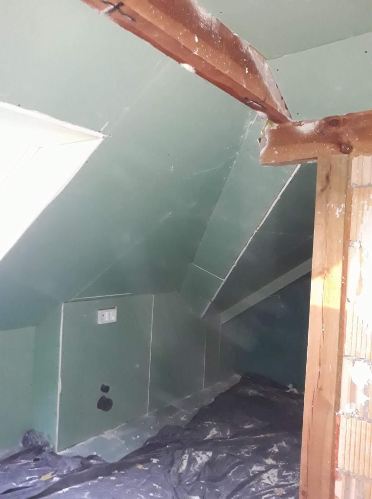 Poddasze w trakcie adaptacji: ściany i skosy wyłożone płytami gipsowo-kartonowymi, widoczne drewniane elementy konstrukcyjne i zabezpieczająca folia malarska.