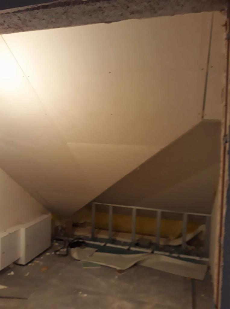 Poddasze z płytami gipsowo-kartonowymi, widoczne metalowe profile i fragment grzejnika, rozrzucone materiały budowlane na podłodze.