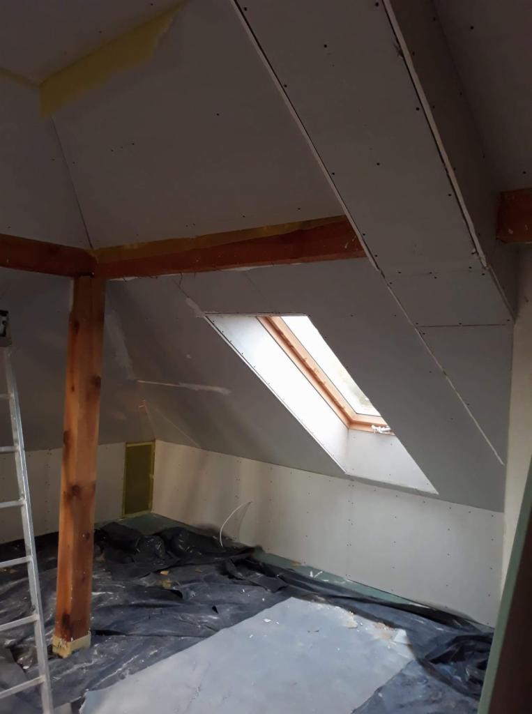Poddasze w trakcie remontu, widoczne płyty gipsowo-kartonowe na skośnym suficie i ścianach, drewniana konstrukcja oraz okno dachowe, podłoga zabezpieczona folią malarską.