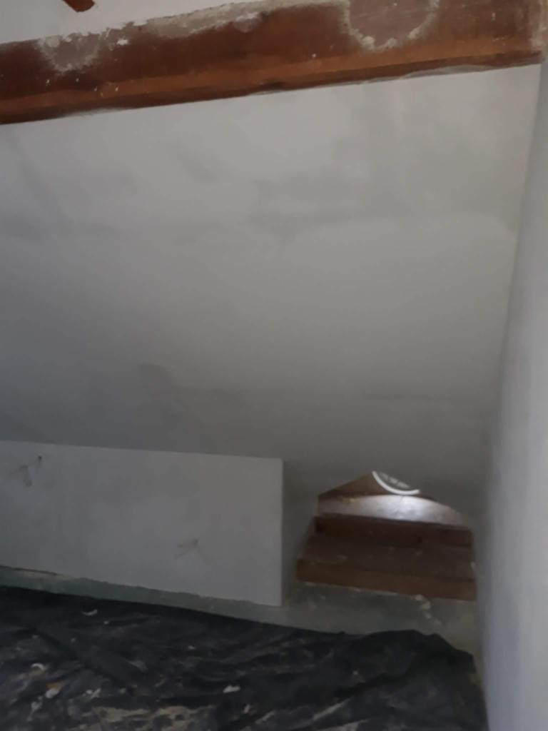 Surowa ściana po wstępnym nałożeniu gładzi, widoczne niedoskonałości, zabezpieczona podłoga folią malarską, fragment drewnianej belki stropowej u góry.