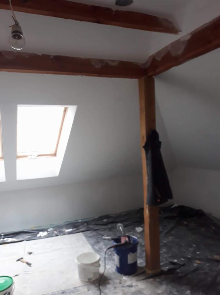 Poddasze w trakcie malowania: widoczne okna dachowe, drewniana konstrukcja, wiadro z farbą, folia ochronna na podłodze i wiszący płaszcz.