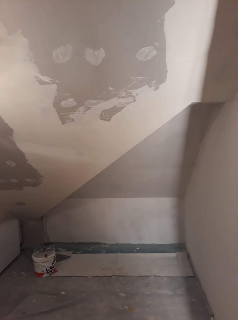 Poddasze w trakcie szpachlowania, widoczne ślady po łataniu dziur na skośnej ścianie, wiadro z masą szpachlową stoi przy ścianie.