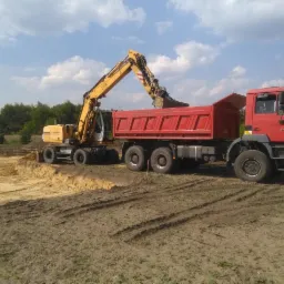 Żółta koparka kołowa Liebherr ładuje urobek do czerwonej wywrotki MAN na placu budowy, widoczne ślady opon na ziemi i wykopane fundamenty w tle.