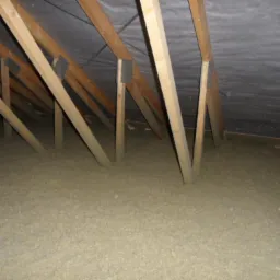 Poddasze z drewnianą konstrukcją krokwiową i warstwą jasnej izolacji termicznej, widok z dołu, ciemne poszycie dachu.