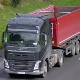 Czarno-szara ciężarówka Volvo z czerwoną naczepą typu wywrotka, widoczna z góry na asfalcie drogi, z zielonym poboczem w tle.