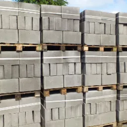 Szare bloczki betonowe ułożone na paletach, zabezpieczone białym sznurkiem, gotowe do transportu. Widok na zewnątrz, w tle zieleń drzew.