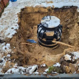 Czarny zbiornik z pokrywą, częściowo przysypany śniegiem, umieszczony w wykopie w ziemi, widoczne narzędzia i fragment butów robotnika.