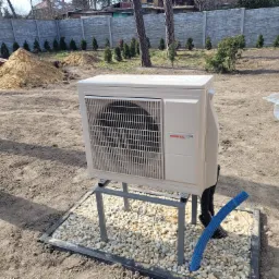 Zewnętrzna jednostka klimatyzacyjna Atlantic R32 zamontowana na metalowym stelażu na żwirowym podłożu, z widocznym niebieskim karbowanym przewodem, w tle sterta ziemi i betonowe ogrodzenie.