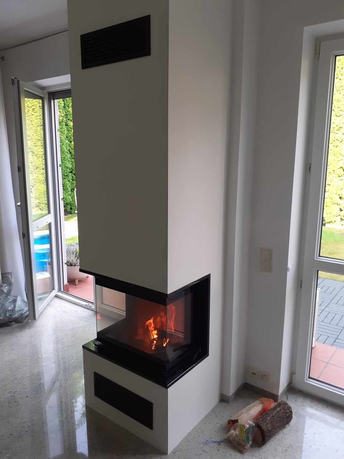 Nowoczesny narożny wkład kominkowy z paleniskiem, obudowany białą konstrukcją z czarnymi akcentami, widoczny przez szklaną szybę. Na podłodze przygotowane drewno do palenia. W tle widoczne okno...