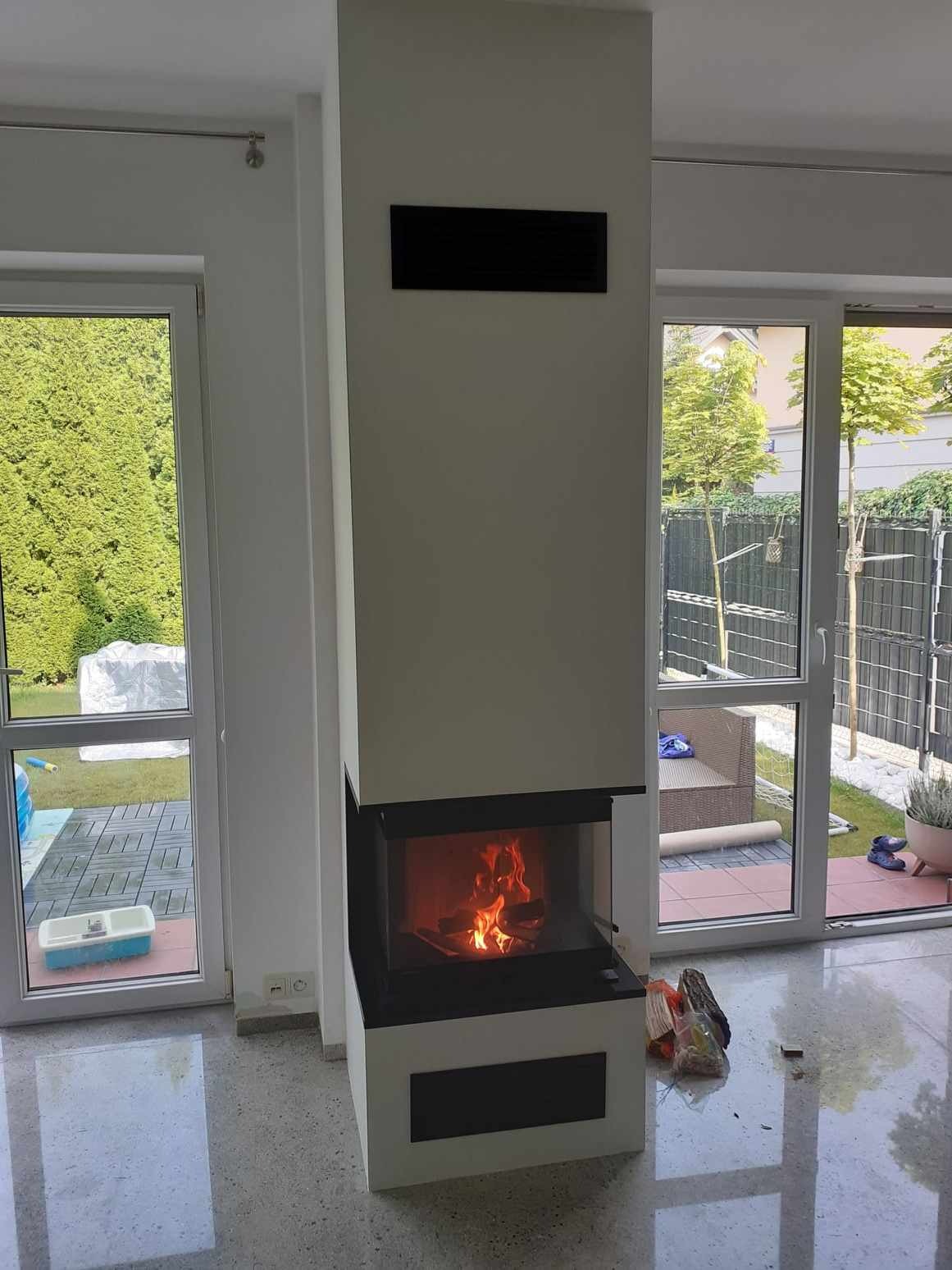 Nowoczesny, narożny wkład kominkowy z paleniskiem, zabudowany w białej obudowie z czarnymi akcentami, widok z boku na płomienie, z widokiem na ogród przez okno tarasowe.