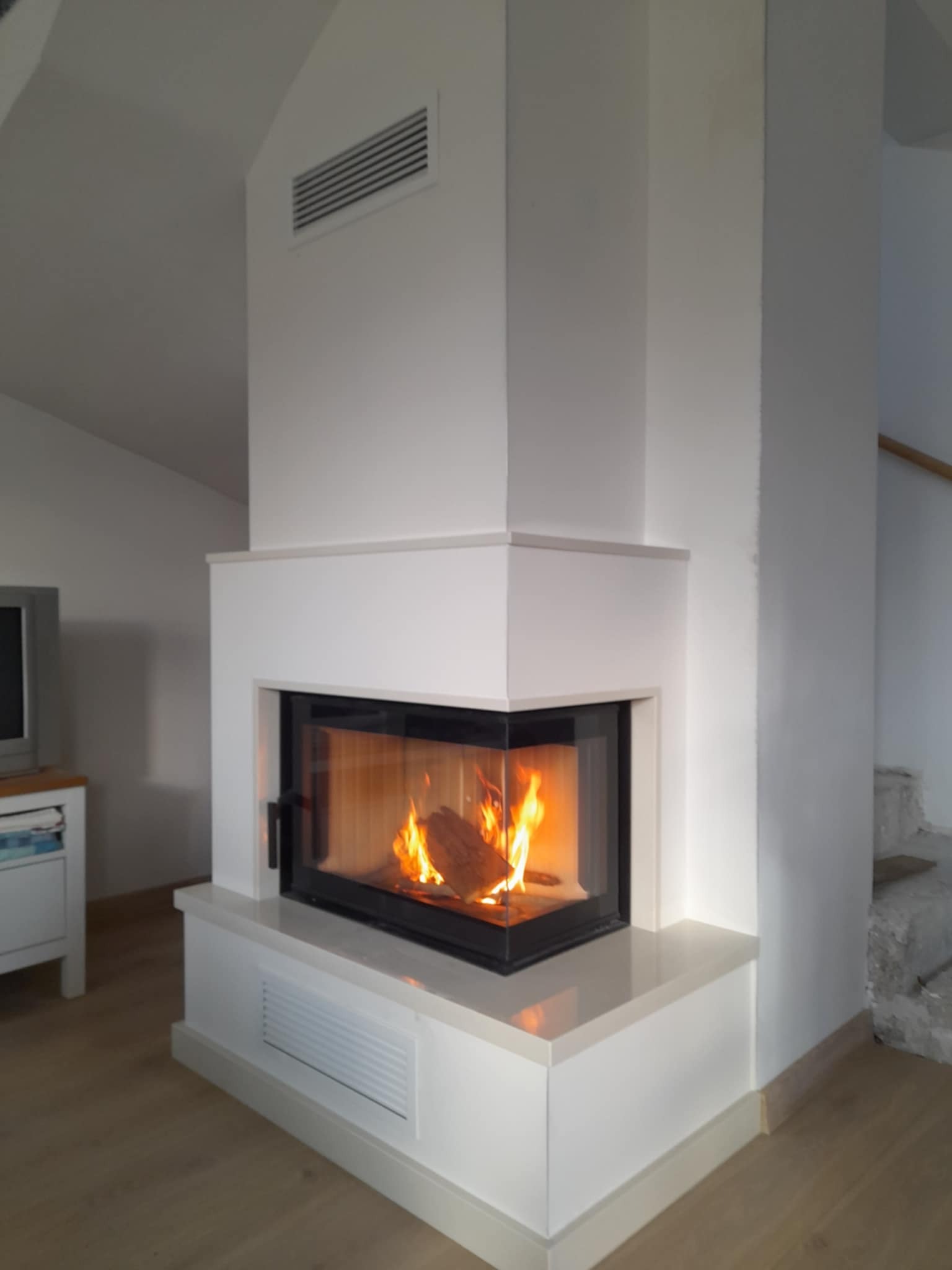 Nowoczesny, narożny wkład kominkowy z czarną ramą i płomieniami, obudowany białą konstrukcją z wentylacją, w jasnym wnętrzu.
