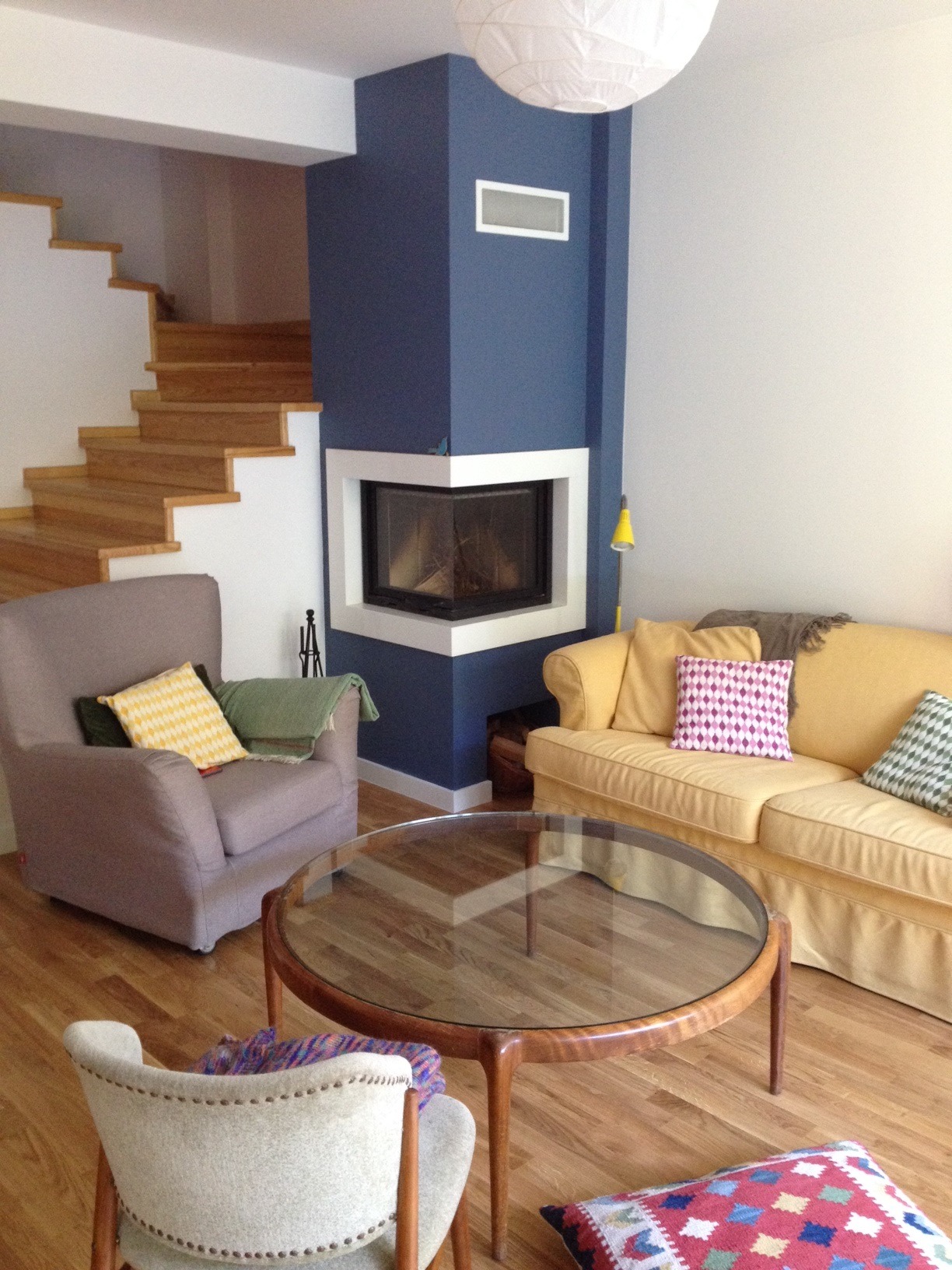 Salon z narożnym wkładem kominkowym obudowanym na granatowo, drewniane schody i parkiet, żółta sofa i szary fotel, okrągły stolik kawowy ze szklanym blatem.