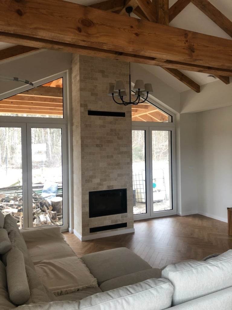 Salon z wyeksponowanym ceglanym obudowanym paleniskiem, drewnianą konstrukcją dachu i parkietem ułożonym w jodełkę, widok zza jasnej sofy