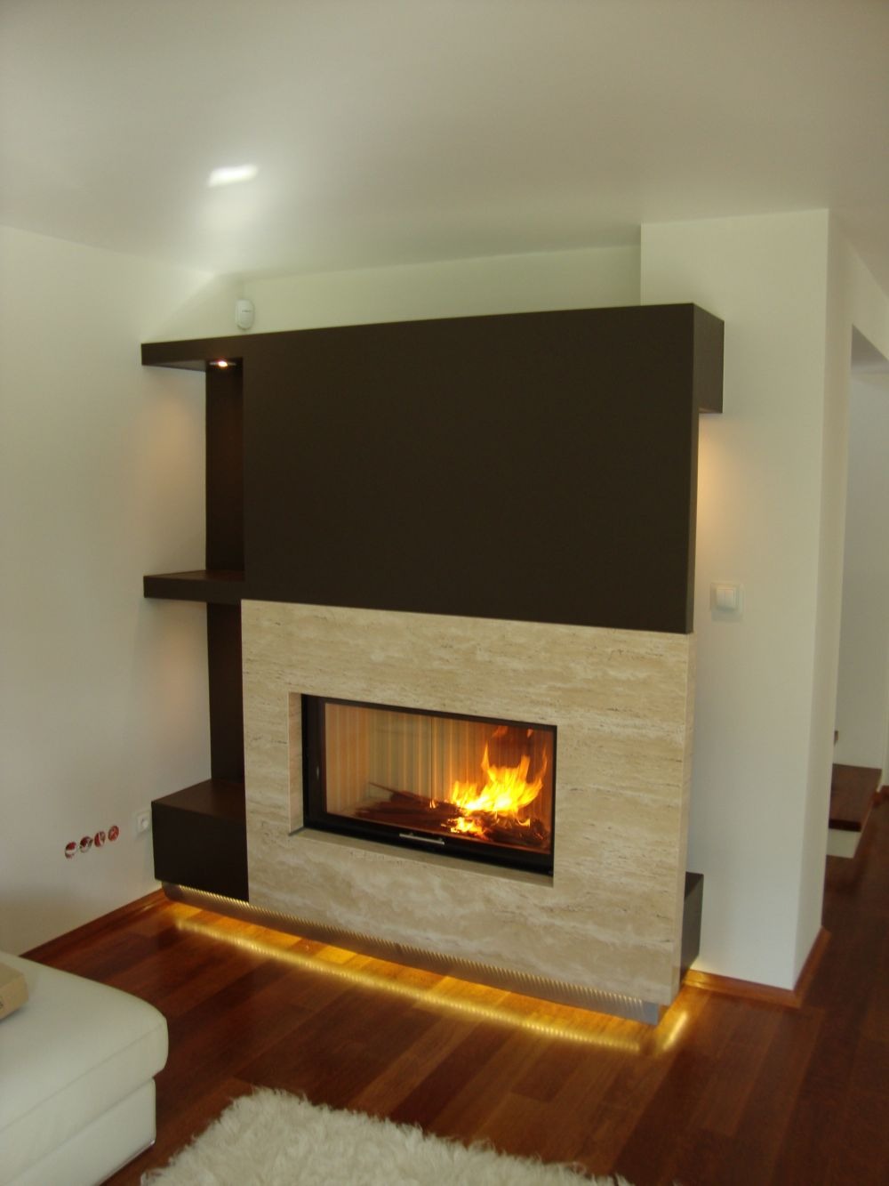 Nowoczesny kominek z obudową z trawertynu, z czarnymi elementami i podświetleniem LED u podstawy, płomienie w palenisku, drewniana podłoga i fragment białego dywanu.