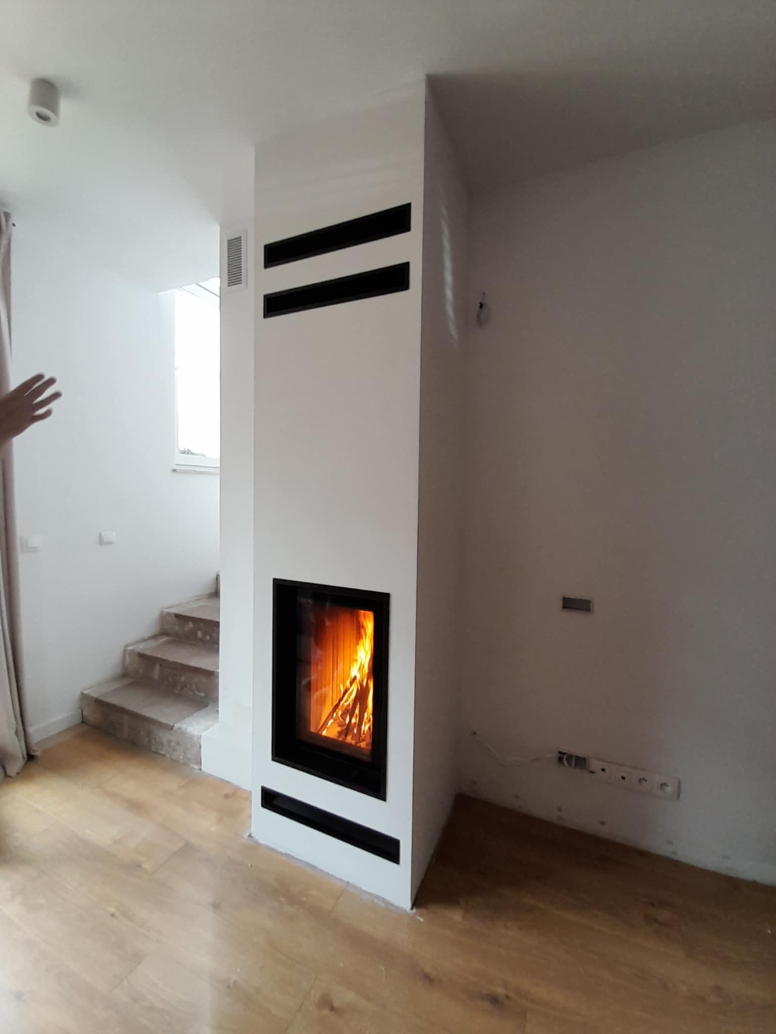 Biały, nowoczesny piec z czarnym obramowaniem paleniska i poziomymi wlotami powietrza, płomienie widoczne za szybą, drewniana podłoga, fragment schodów i ręka w kadrze.