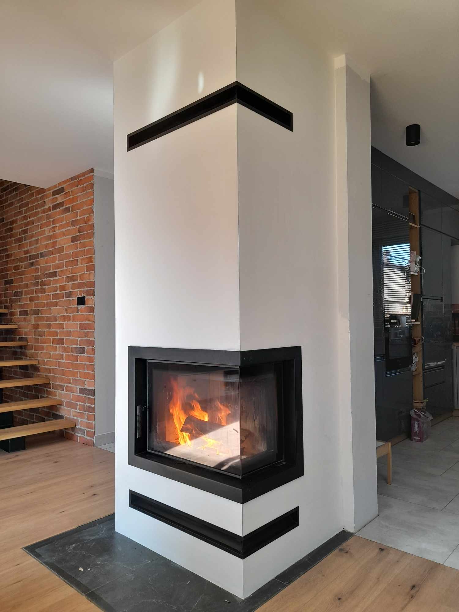 Nowoczesny narożny wkład kominkowy z czarną ramą, obudowany białą płytą, z widocznym płomieniem, na tle ceglanej ściany i drewnianych schodów.