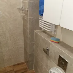 Nowoczesna łazienka z beżowymi płytkami imitującymi marmur, drewnopodobnymi panelami na podłodze, grzejnikiem drabinkowym z niebieskim ręcznikiem i toaletą z chromowanym przyciskiem spłukiwania.