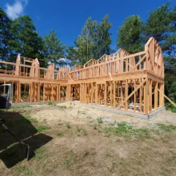 Konstrukcja drewniana domu w stanie surowym otwartym, widoczne fundamenty i szkielet ścian, tło stanowią drzewa i błękitne niebo.