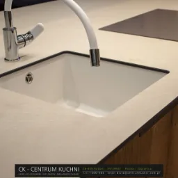 Beżowy blat kuchenny z wbudowanym białym zlewem, chromowaną baterią z elastyczną wylewką i dozownikiem mydła, płyta indukcyjna w tle.