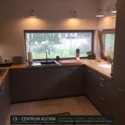 Szara kuchnia z drewnianym blatem pod oknem z żaluzjami, widoczne oświetlenie punktowe i sprzęt AGD, minimalistyczny design.