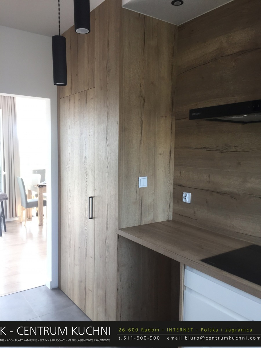 Nowoczesna kuchnia z drewnianymi frontami szafek i blatem, czarne lampy wiszące, widok na jadalnię w tle.