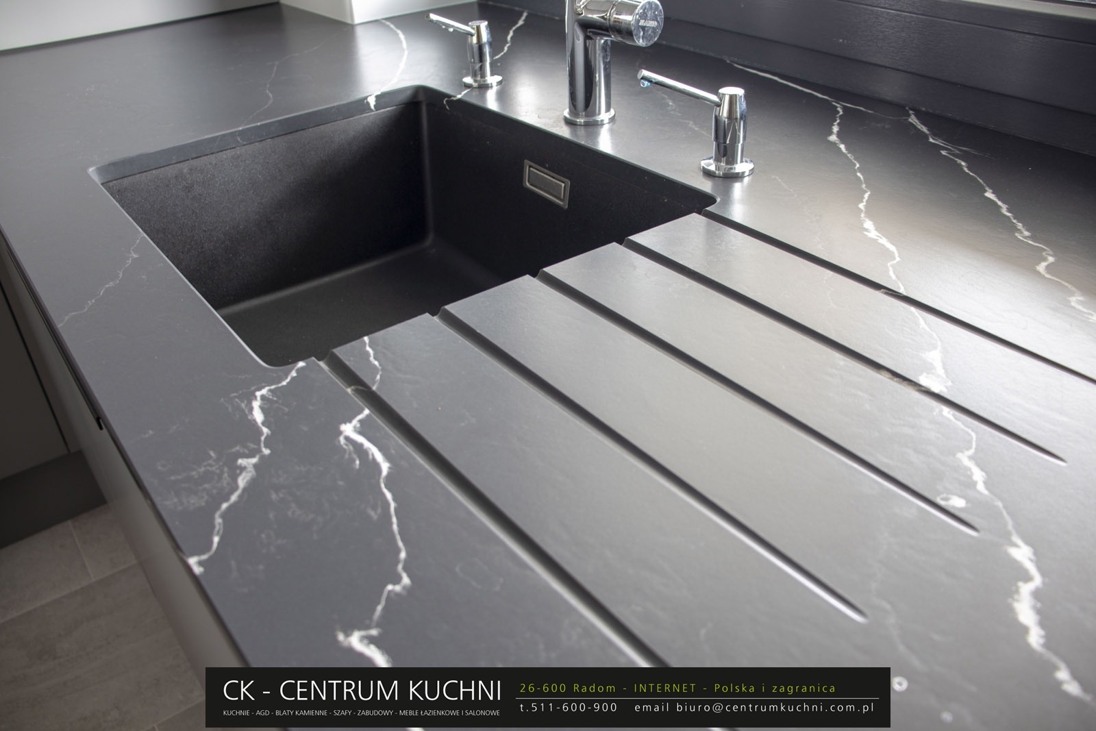 Czarny, kamienny blat kuchenny z wbudowanym zlewem i ociekaczem, bateria w kolorze chromu i dozowniki na płyn, widoczne marmurkowe żyły na blacie.
