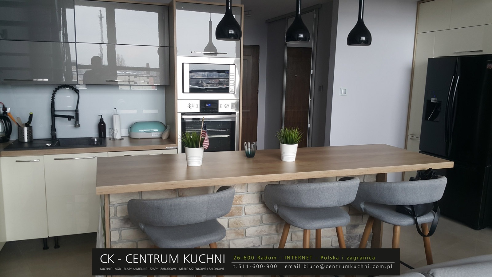 Nowoczesna kuchnia z wyspą, fronty szafek w odcieniach szarości i beżu, blat z jasnego drewna, wyspa wykończona cegłą, trzy szare krzesła barowe, czarne lampy wiszące.