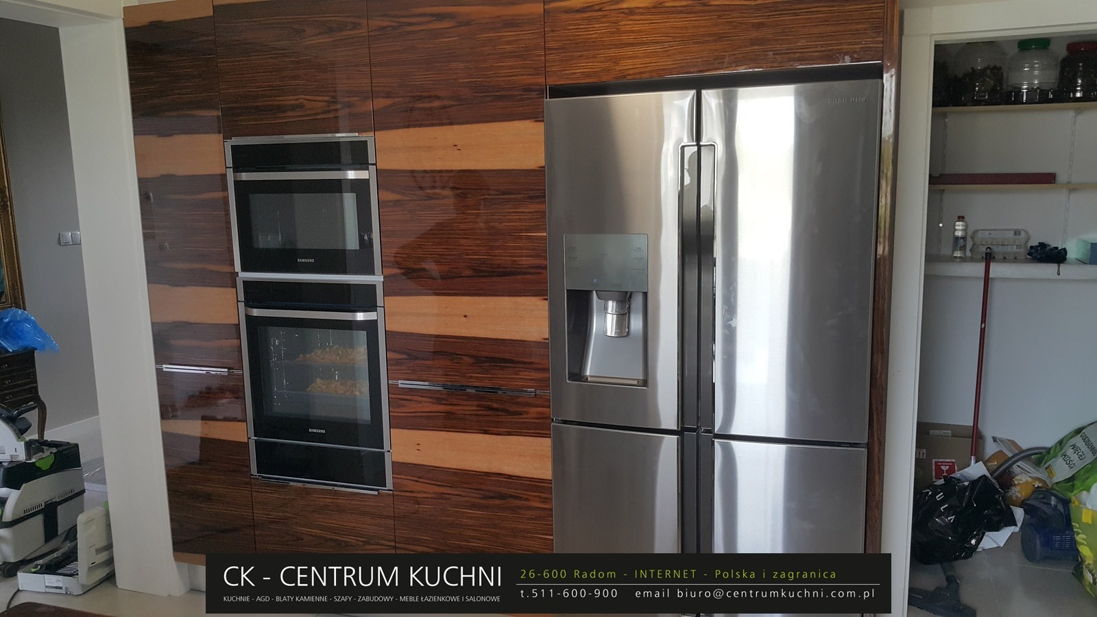 Nowoczesna zabudowa kuchenna z fornirem o egzotycznym wzorze, zintegrowana z dwoma piekarnikami Samsung i lodówką side-by-side, widoczny fragment spiżarni obok.