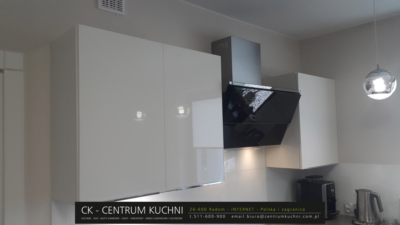 Białe, lakierowane szafki kuchenne wiszące z czarnym okapem i okrągłą lampą wiszącą, widoczne ekspres do kawy i czajnik elektryczny na blacie.