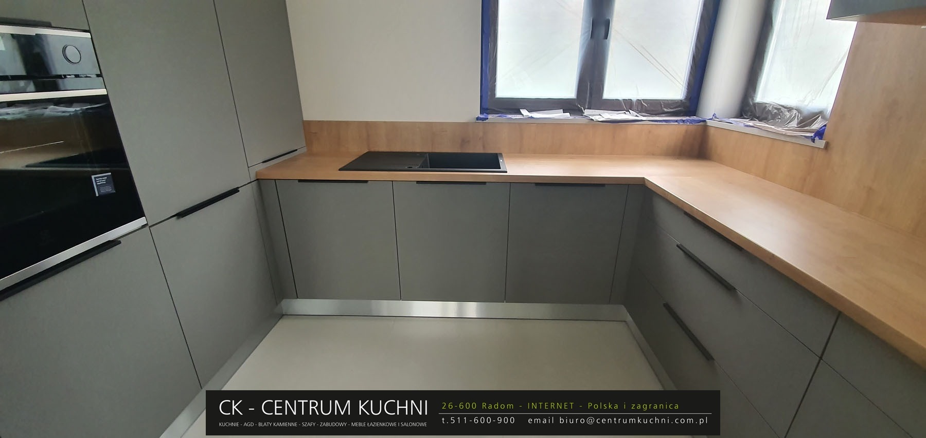 Szara zabudowa kuchenna z drewnianym blatem i czarnym zlewem, okno zabezpieczone folią malarską, nowoczesny minimalizm.